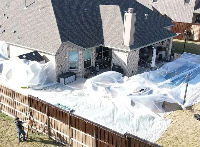 Aerial view of a backyard covered in plastic sheeting while a crew member stains the fence